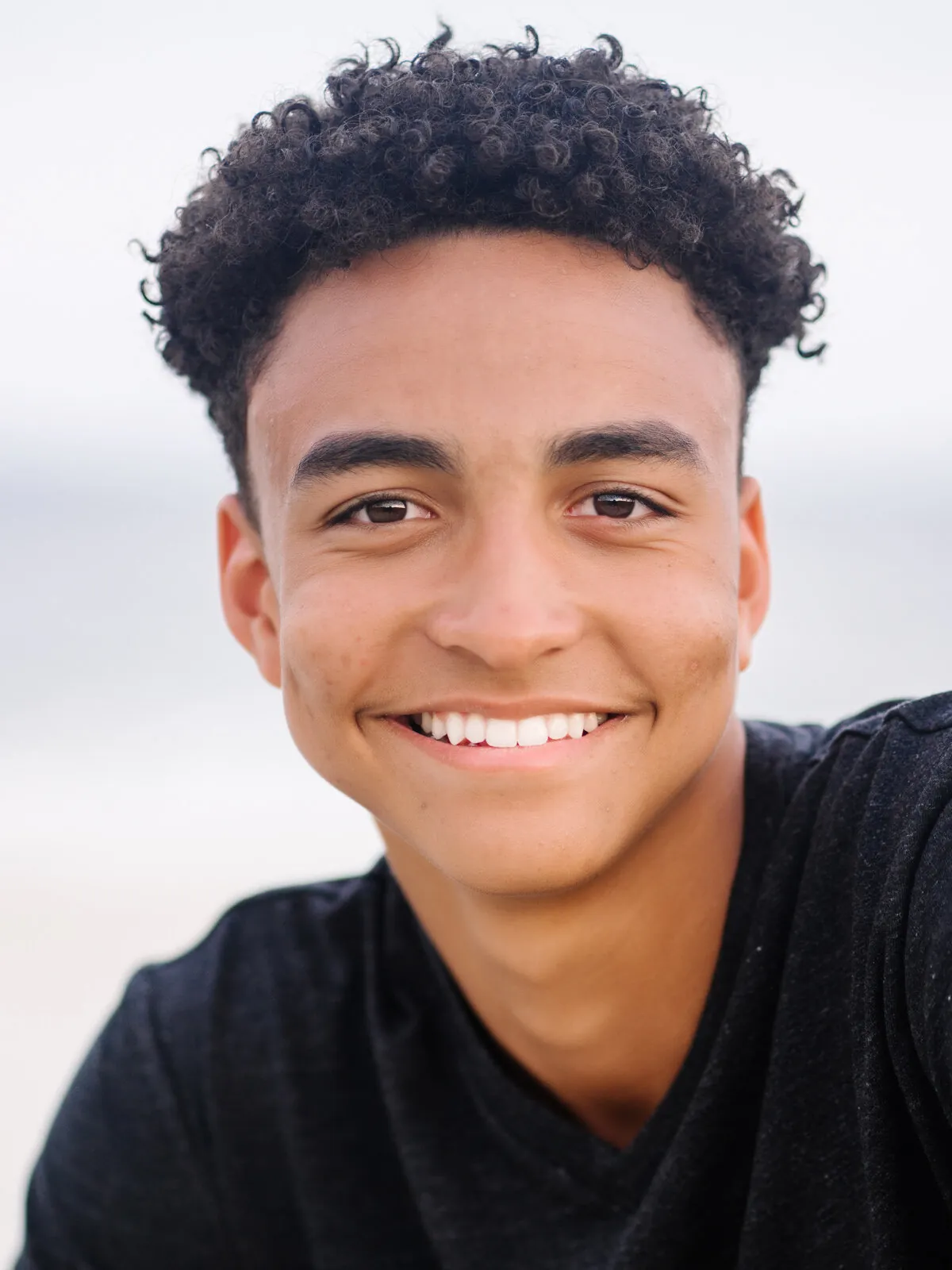 Professional headshot of a smiling teenager with curly hair in a black t-shirt, outdoors by Myrtle Beach headshot photographer Pasha Belman
