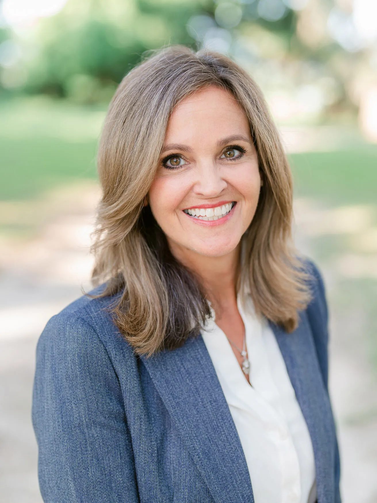 Professional headshot of a smiling woman with shoulder-length hair in a blue-gray blazer and white blouse, outdoors under live oaks by Myrtle Beach headshot photographer Pasha Belman