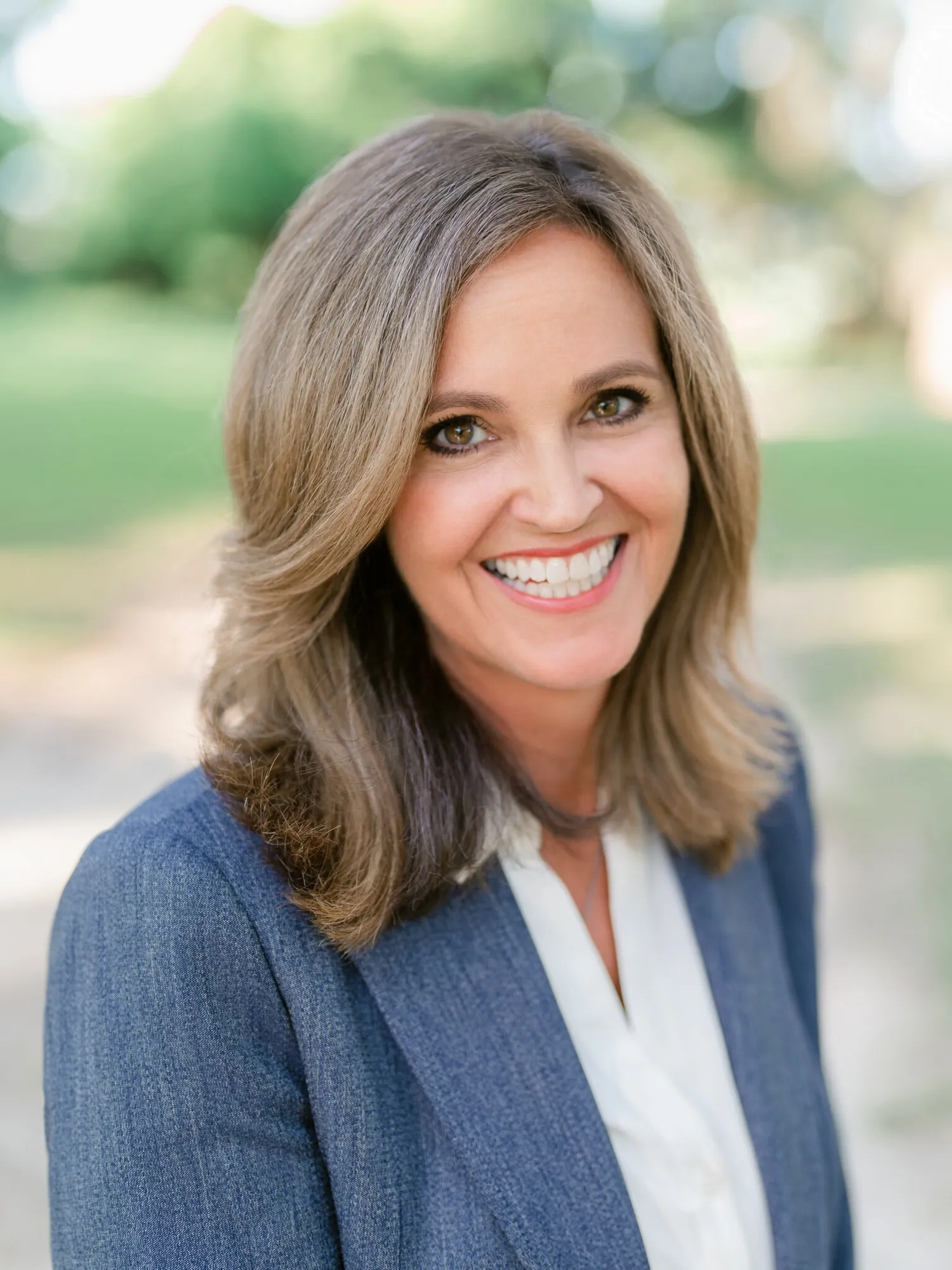 Professional headshot of a woman with shoulder-length hair in a blue-gray blazer and white blouse smiling warmly, outdoors by Myrtle Beach headshot photographer Pasha Belman