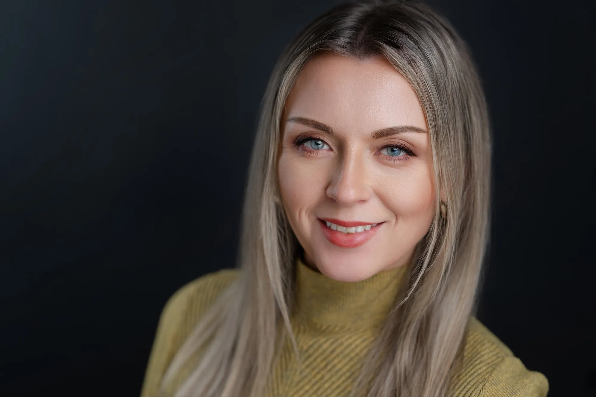Professional headshot of a smiling woman with long hair in a mustard turtleneck against a dark backdrop by Myrtle Beach headshot photographer Pasha Belman