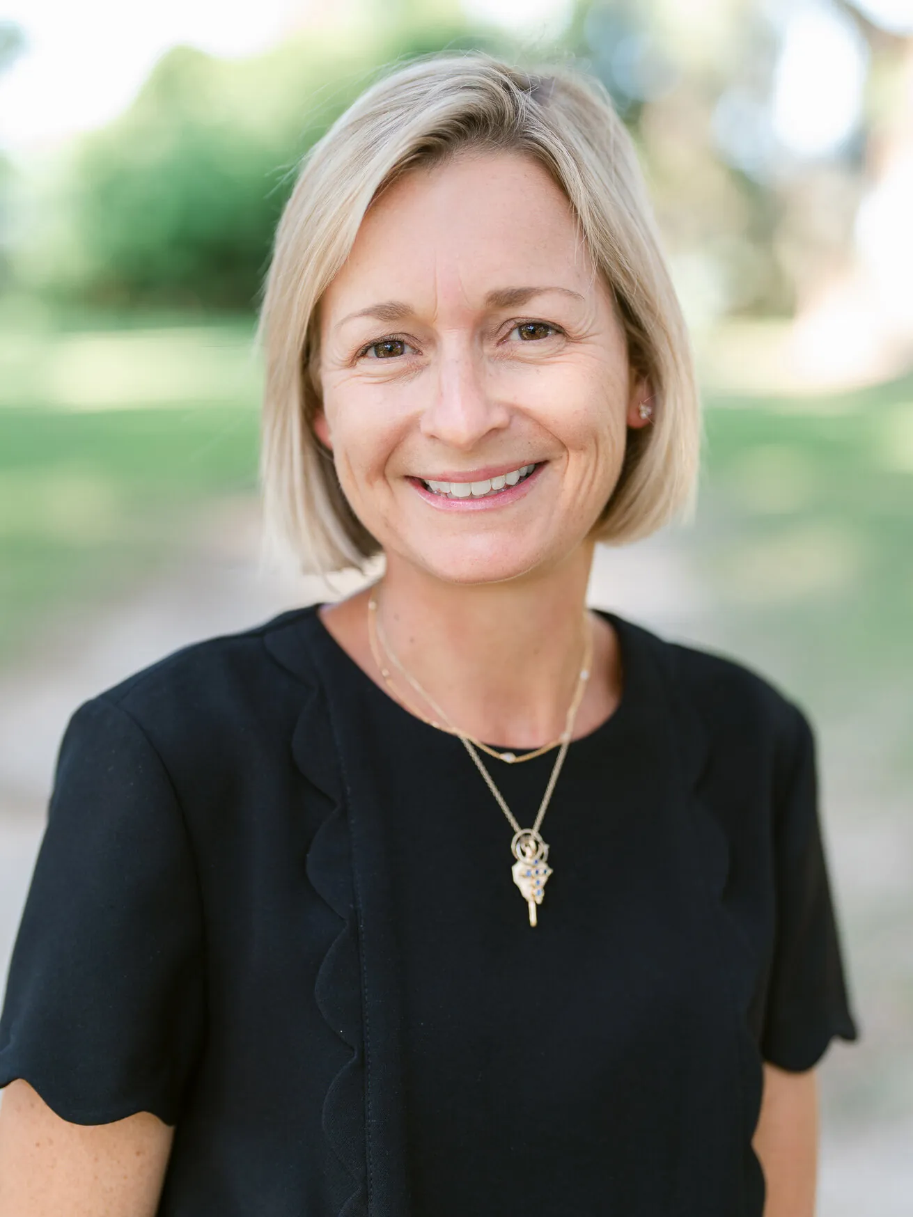 Professional headshot of a woman with a short blonde bob in a black top and pendant necklace, outdoors by Myrtle Beach headshot photographer Pasha Belman
