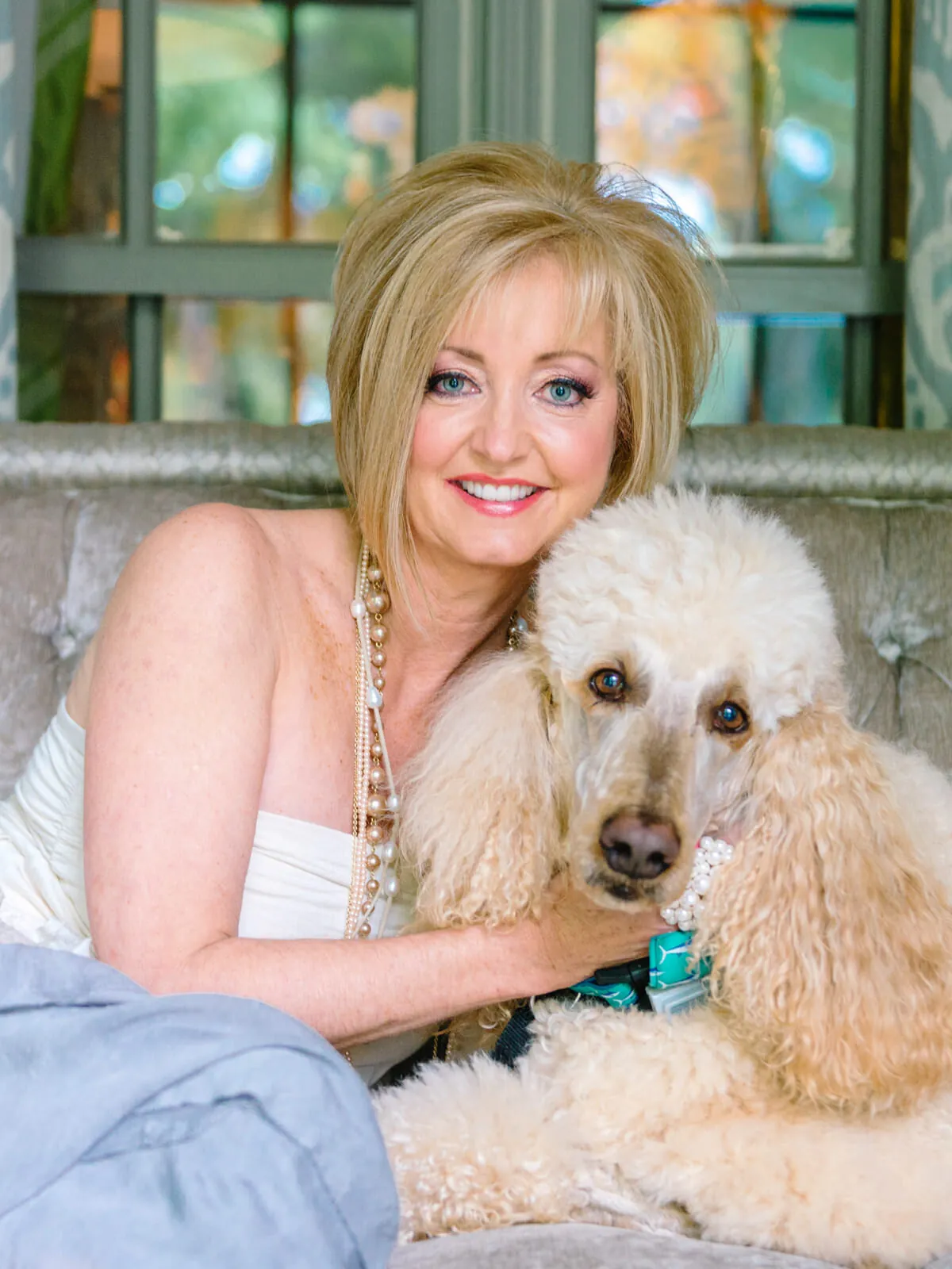 Professional headshot of a smiling woman with blonde hair holding her white poodle on a chaise by a window by Myrtle Beach headshot photographer Pasha Belman