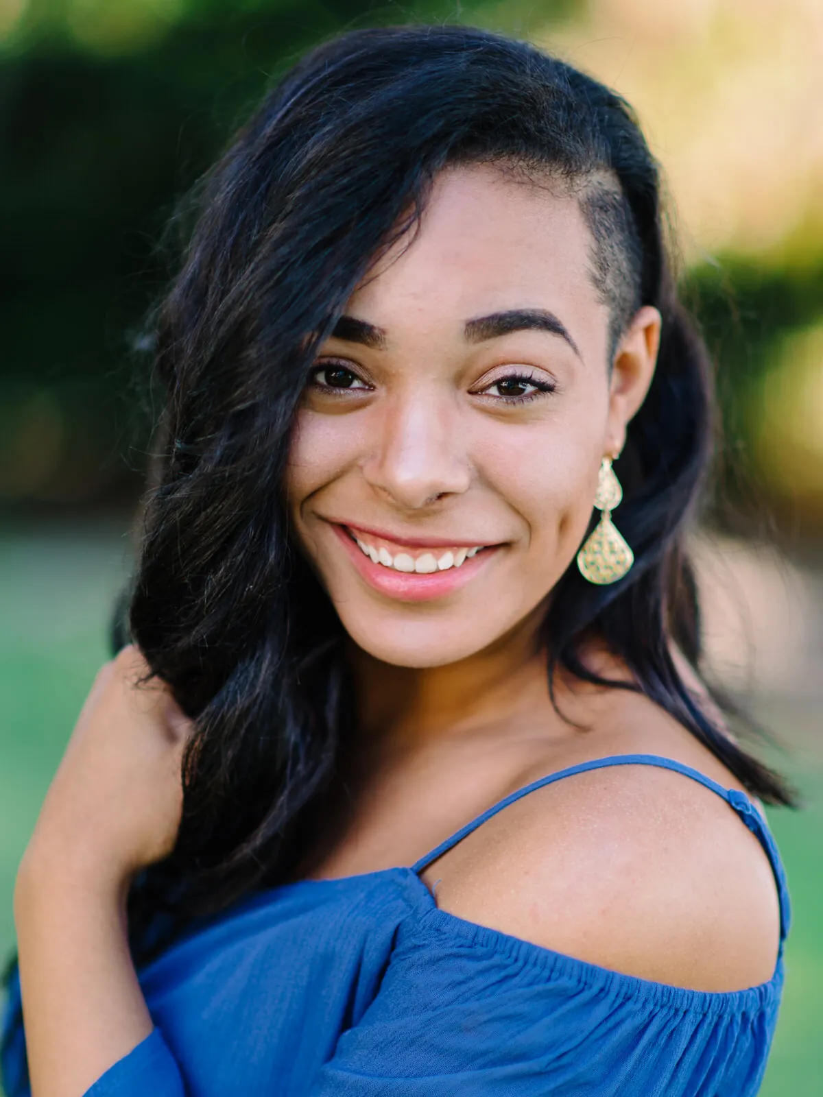 Professional headshot of a smiling woman with black wavy hair and gold earrings in a blue off-shoulder top by Myrtle Beach headshot photographer Pasha Belman