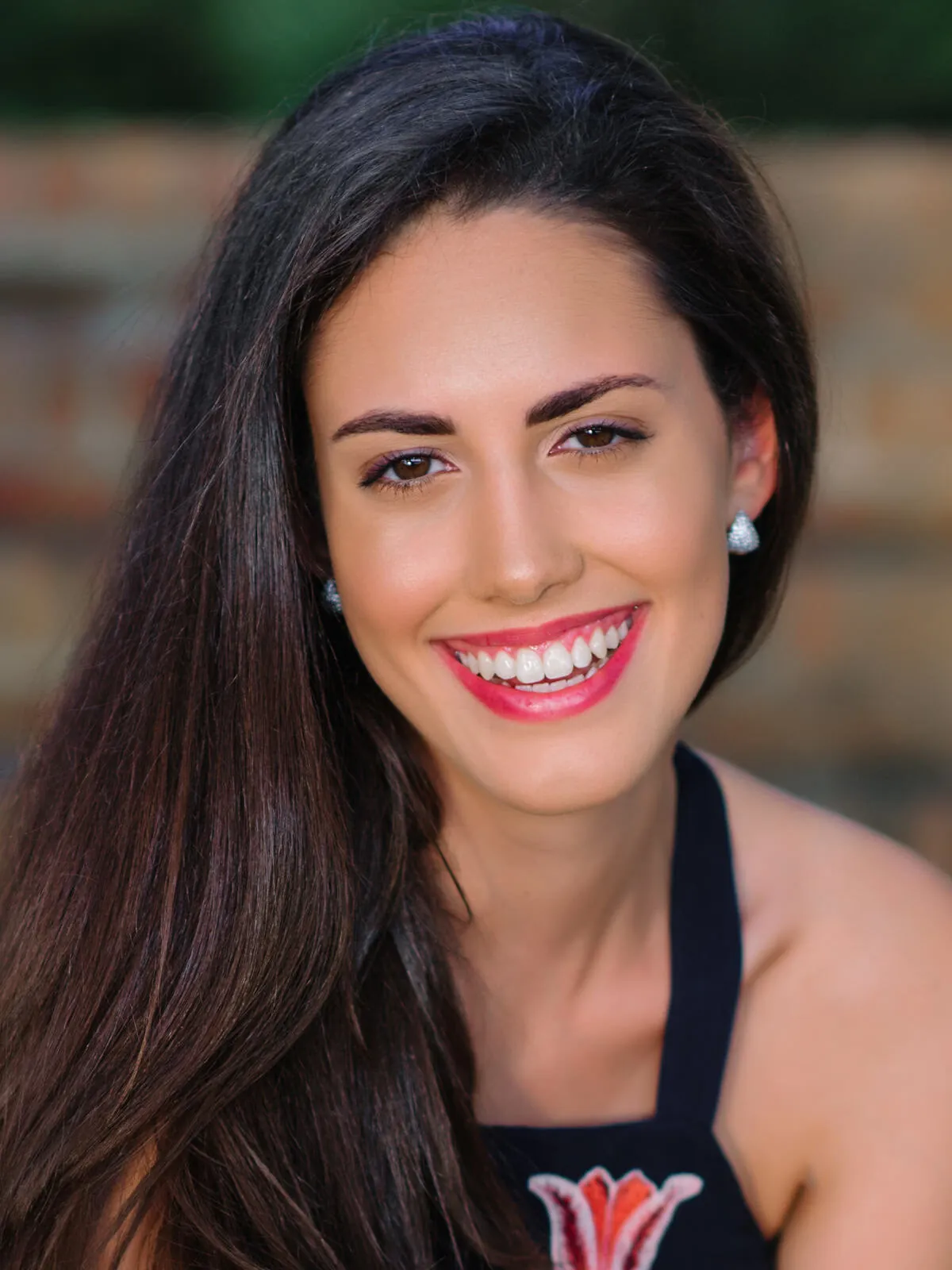 Professional headshot of a smiling woman with long dark hair and red lipstick in a black top, in front of a brick wall by Myrtle Beach headshot photographer Pasha Belman
