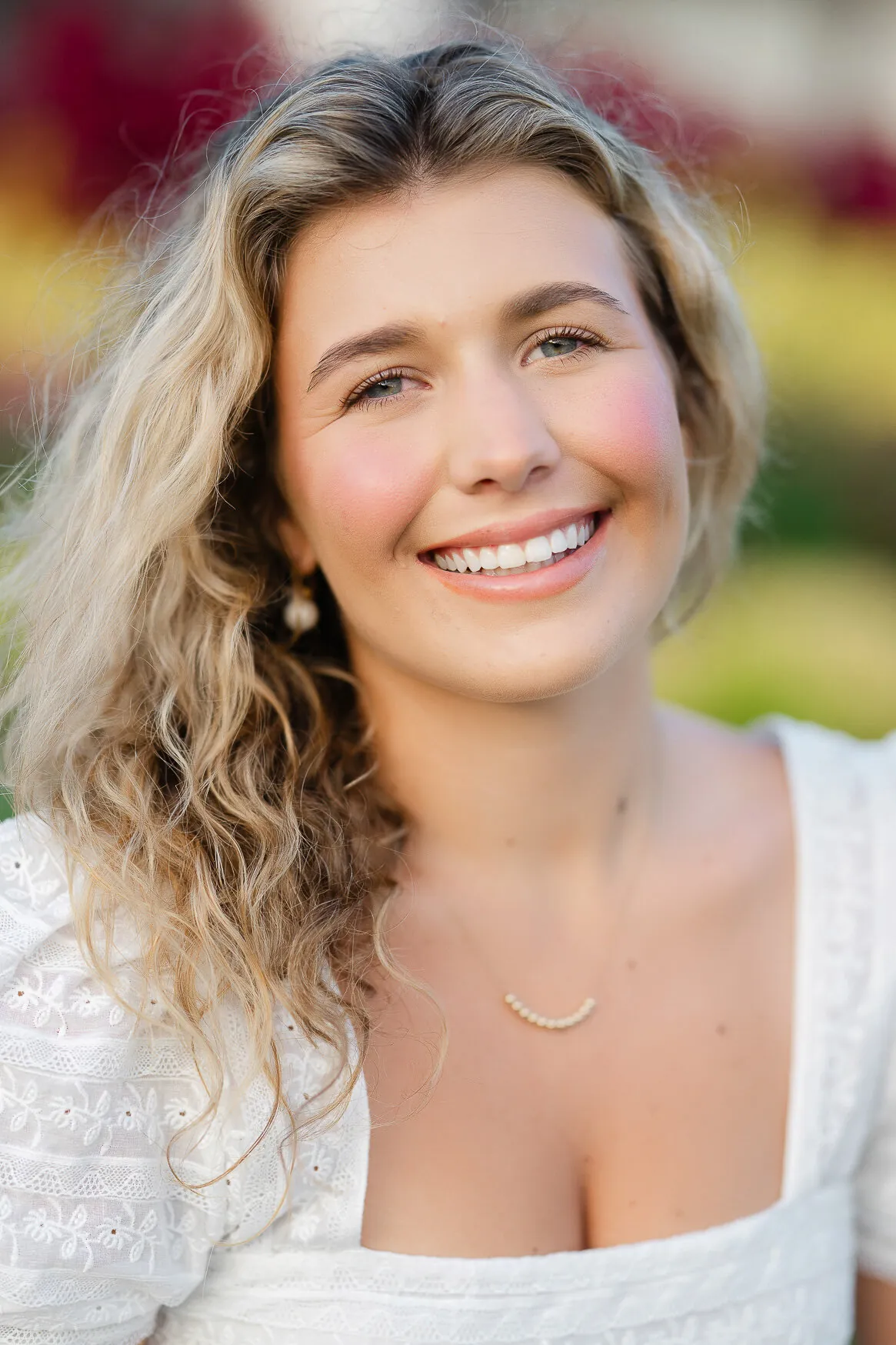 Professional headshot of a young woman with curly blonde hair in a white lace top smiling in front of colorful flowers by Myrtle Beach headshot photographer Pasha Belman