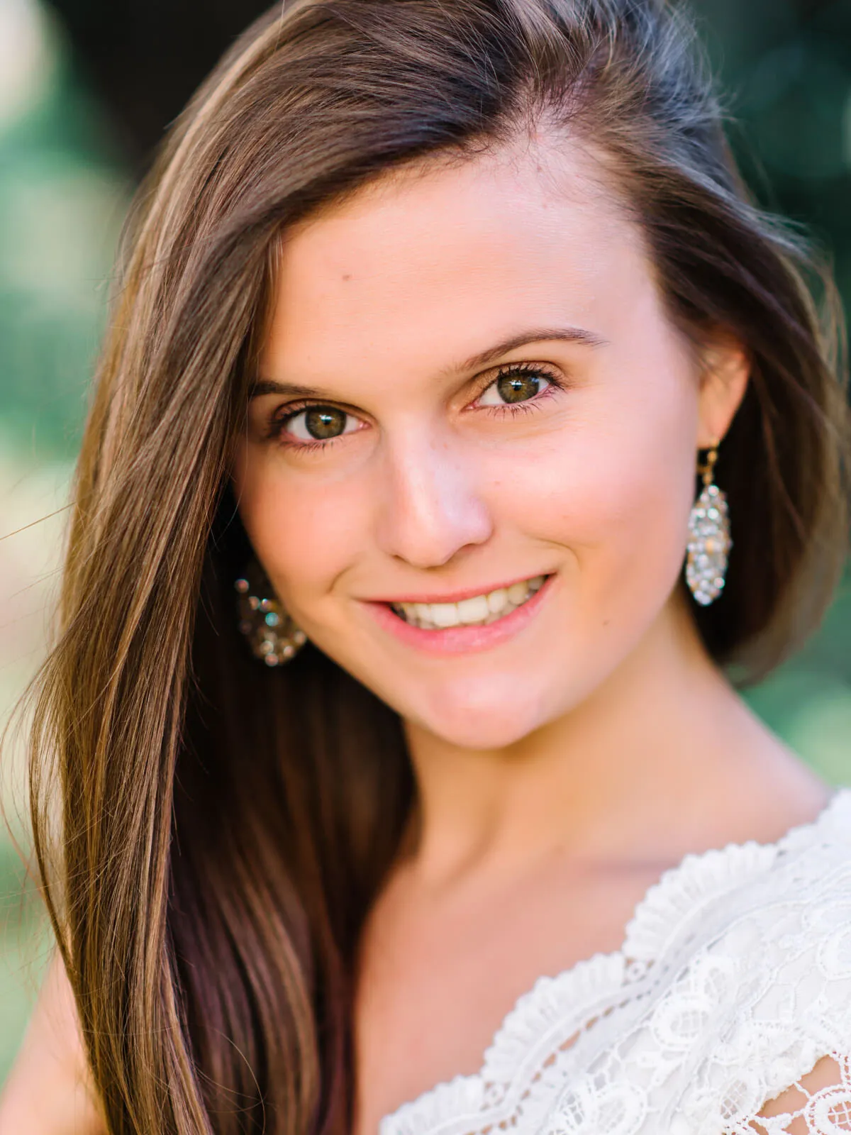 Professional headshot of a smiling young woman with long brown hair, drop earrings and a white lace top by Myrtle Beach headshot photographer Pasha Belman