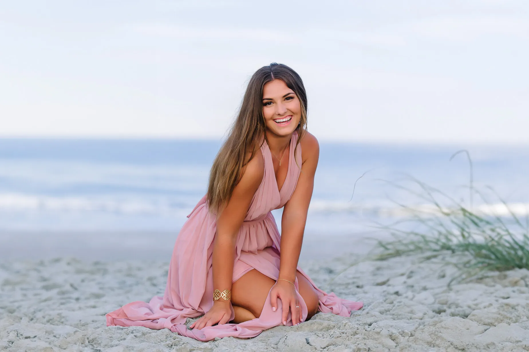 Smiling young woman in a pink dress kneeling on the beach with sea oats behind her, a portrait and personal branding photograph by Myrtle Beach photographer Pasha Belman