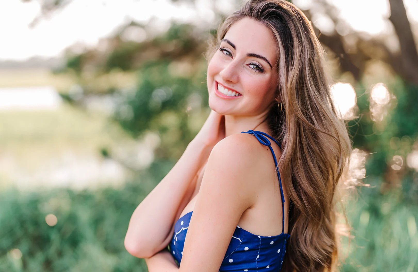 High school senior in a navy polka dot top smiling over her shoulder under oak trees in golden evening light, Myrtle Beach senior photography by Pasha Belman