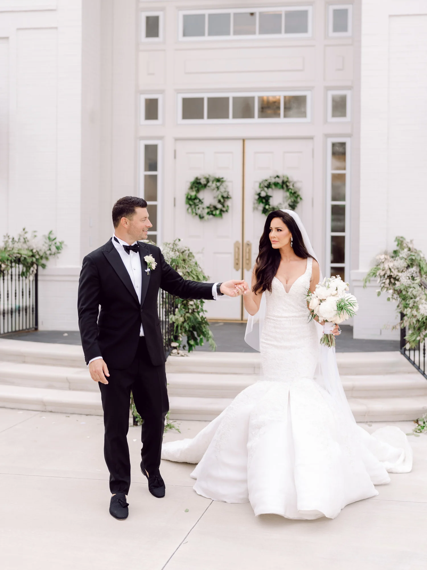Bride and groom holding hands on the steps of a white church with wreaths on the doors, by Myrtle Beach wedding photographer Pasha Belman