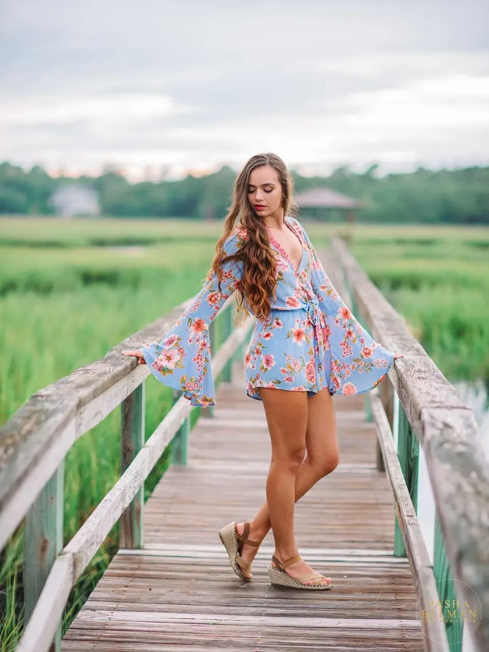 Senior girl in a floral romper and wedge sandals standing on a weathered marsh boardwalk on Pawleys Island