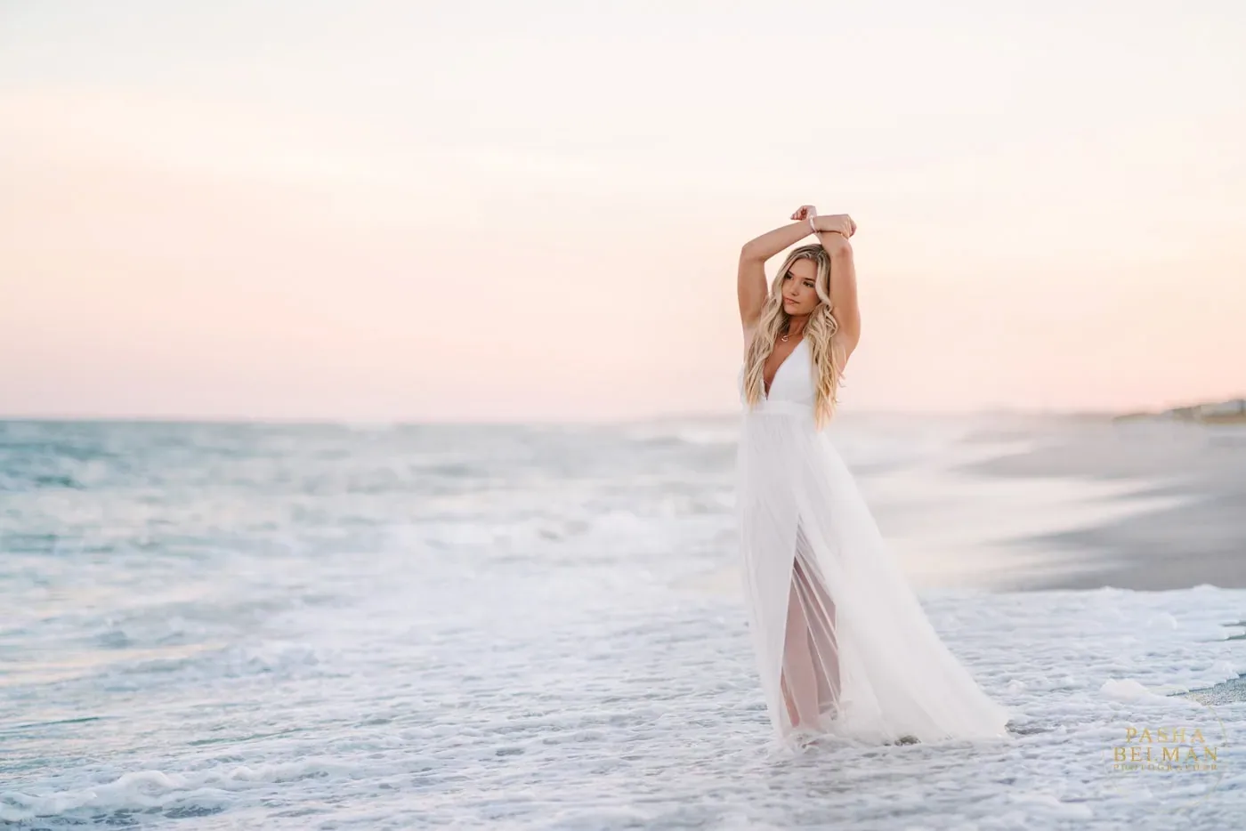 Wide view of a senior girl in a white dress with her arms raised in the surf at sunset on Pawleys Island