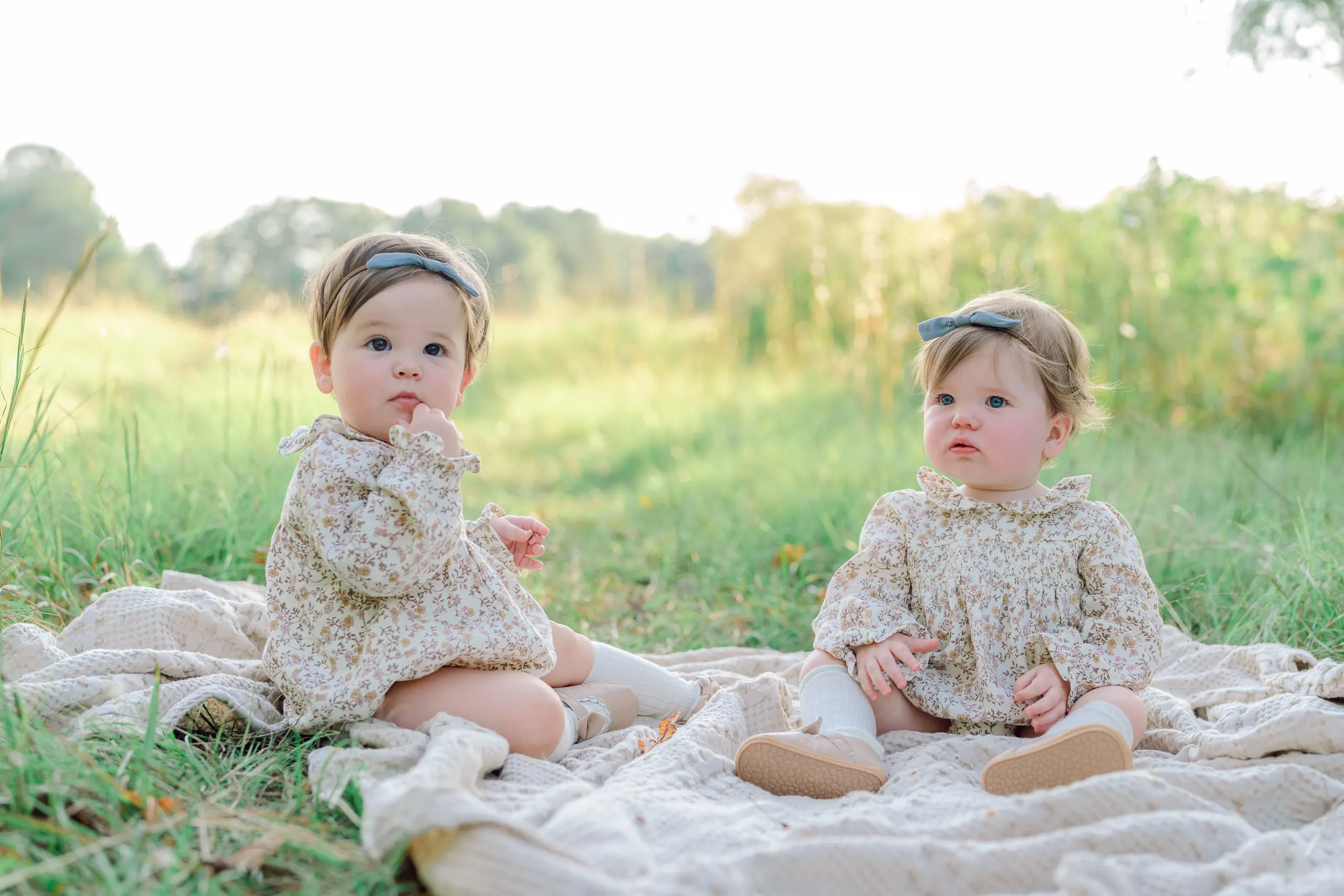 Two babies sitting together on a cream blanket in a green field
