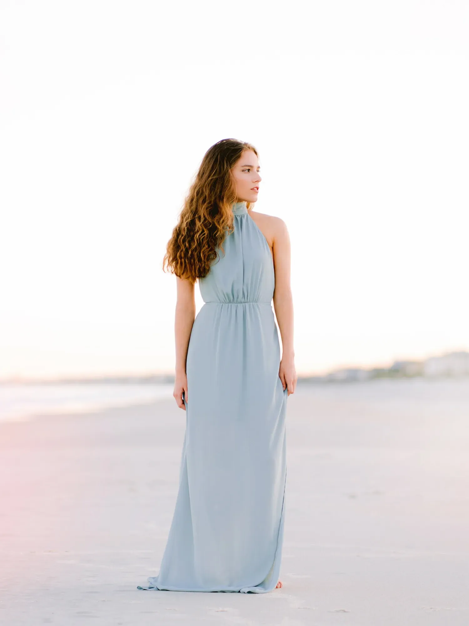 Senior girl in a long blue dress walking along an empty beach at soft sunset