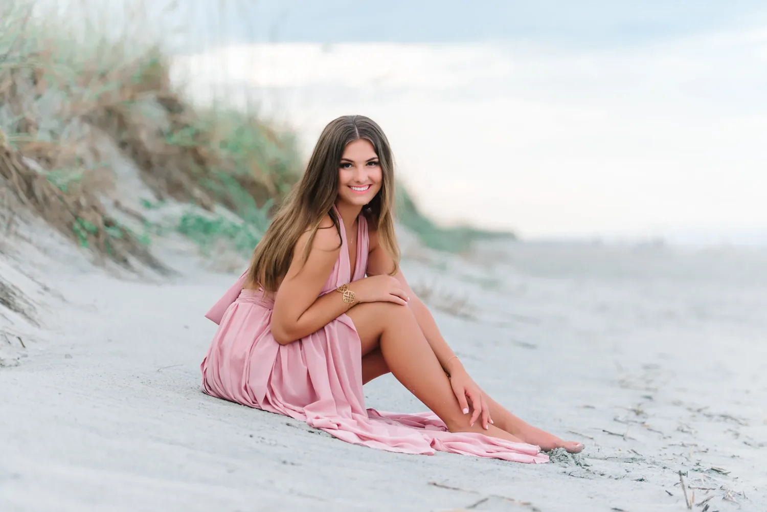 Wide view of a senior girl in a pink dress sitting on a sand dune with sea oats