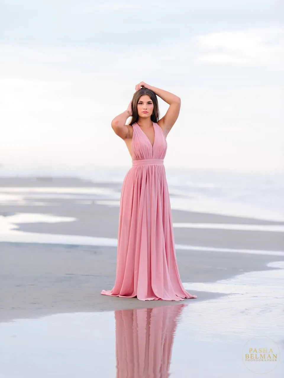 Senior girl in a pink dress with her hands in her hair on reflective wet sand