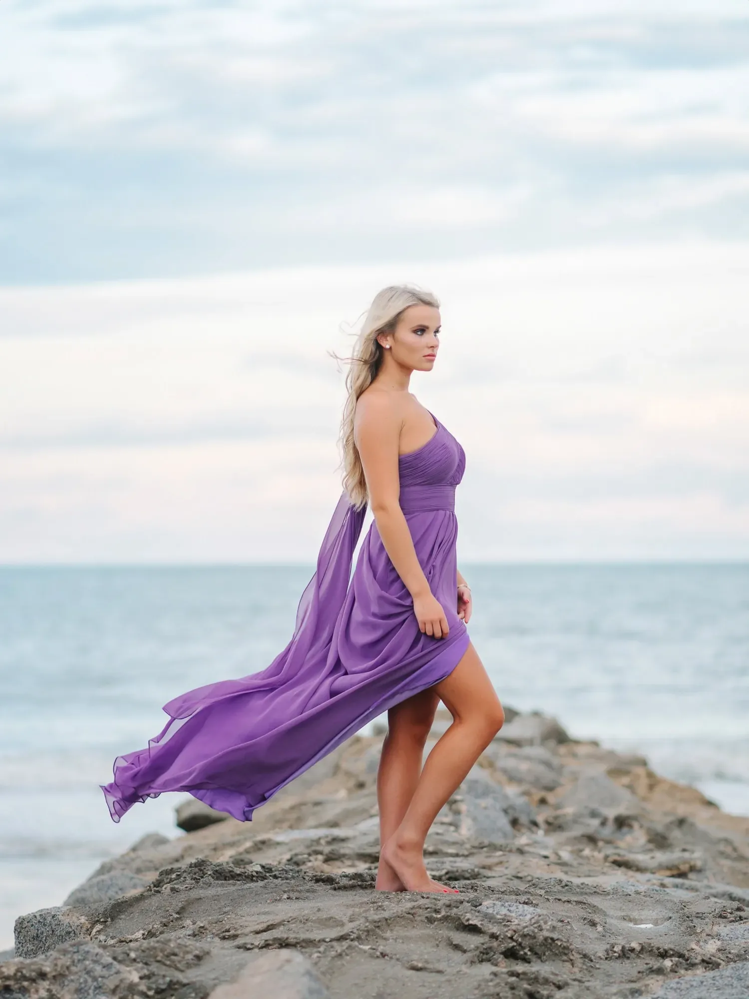 Senior girl in a flowing purple dress posing on coastal rocks above the ocean