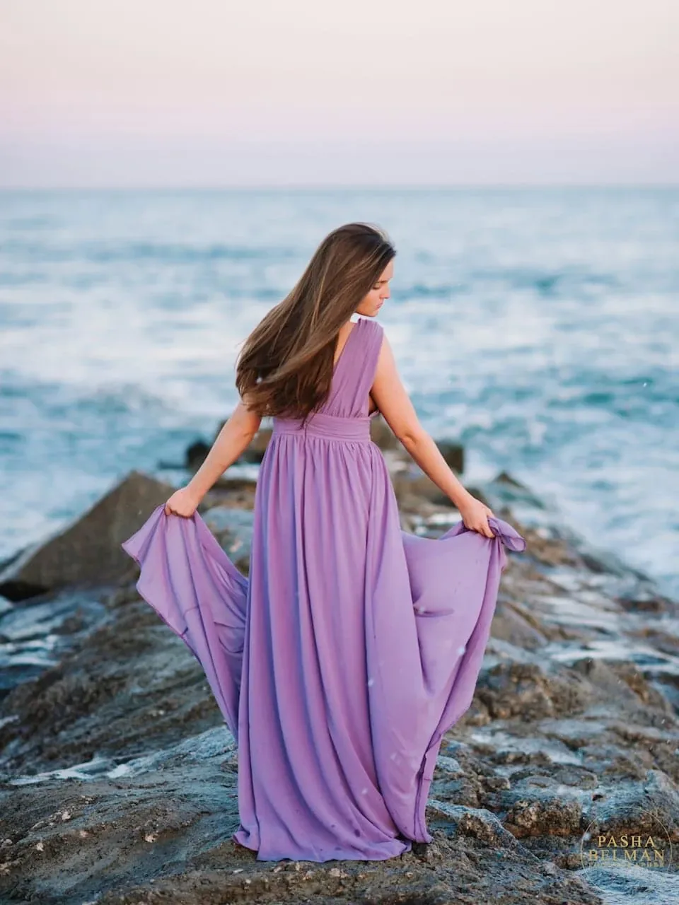 Senior girl in a purple dress holding her skirt and standing on jetty rocks by the sea