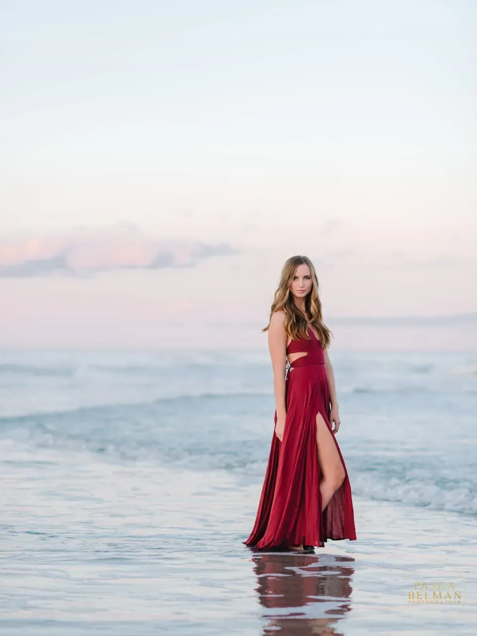 Senior girl in a deep red dress standing in the shallow surf at sunset