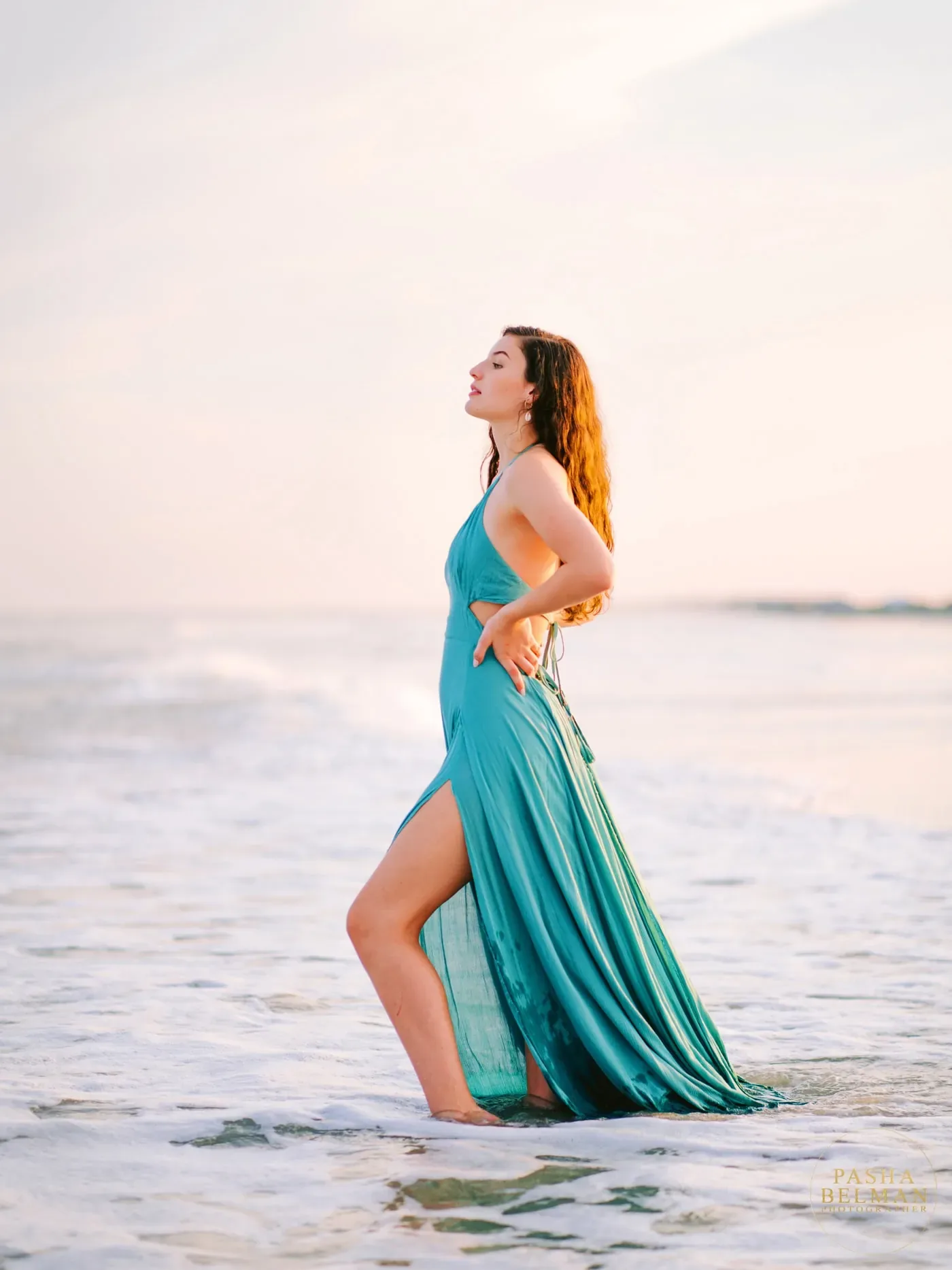 Senior girl in a teal dress standing in the surf with her hand on her hip at sunset