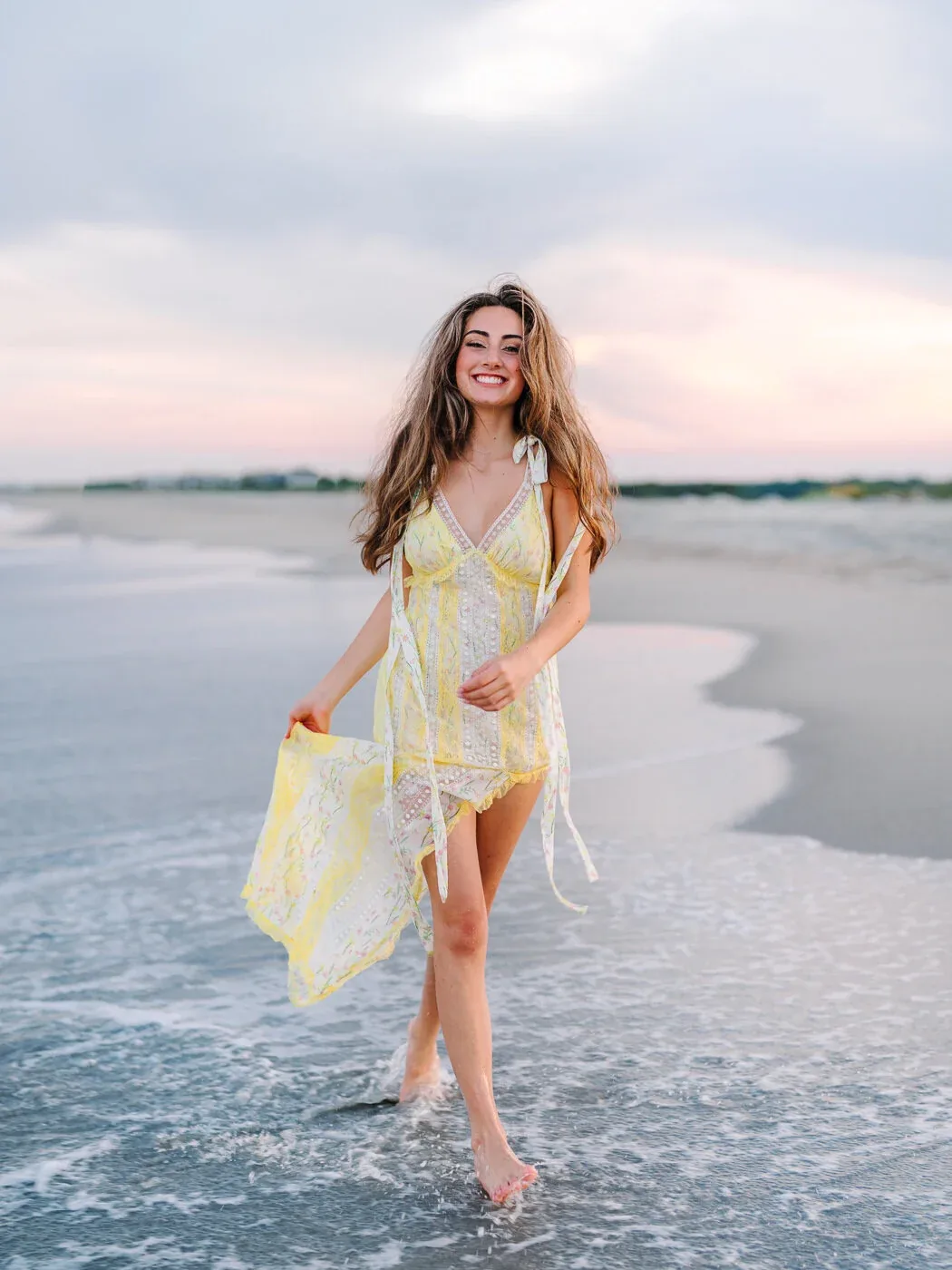 Senior girl in a yellow patterned dress walking through the surf holding her skirt