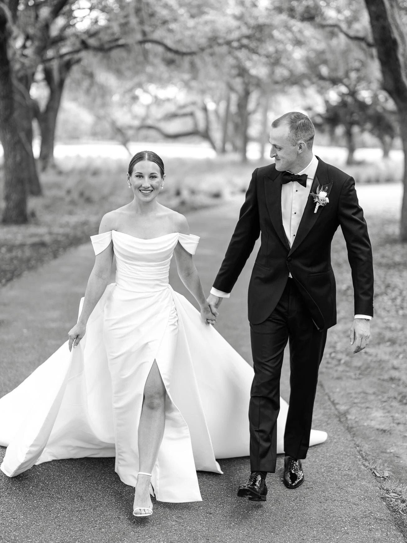 Black and white photo of a bride in an off the shoulder gown and a groom in a tuxedo walking hand in hand under live oak trees, South Carolina wedding