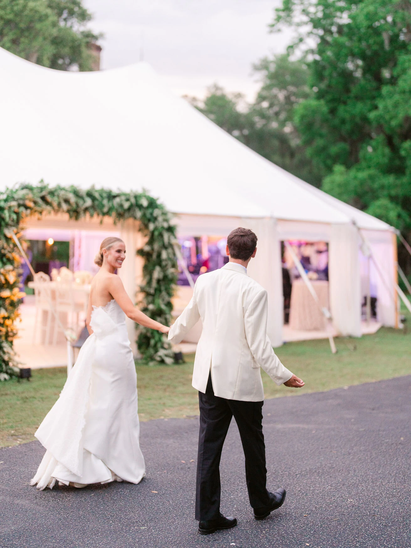 Bride looking back and smiling as she holds the groom's hand while they walk toward their white sailcloth tent reception with a greenery and flower arch, South Carolina wedding